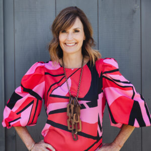 Katie Pulsifer in a vibrant red, pink, and black patterned dress, accessorized with a wooden long necklace, and medium-length hair