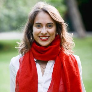 Nina Urman, a life coach with curly blond hair and a noticeable red scarf, wearing a white long sleeve top, set against a serene garden backdrop