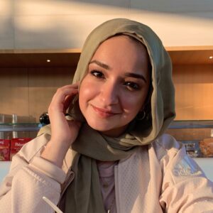 Lama Aboubakr with a radiant smile, outfitted in a white sweater and olive green hijab, amidst a minimalist kitchen setting