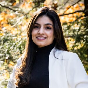 Sukhi Kaur smiling in a black turtleneck and white jacket, with long black hair, set against an autumn backdrop